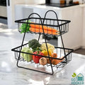GoHooked black metal 2-tier fruit basket with carrots, broccoli, cauliflower, cabbage, peppers, and onions on a marble countertop.