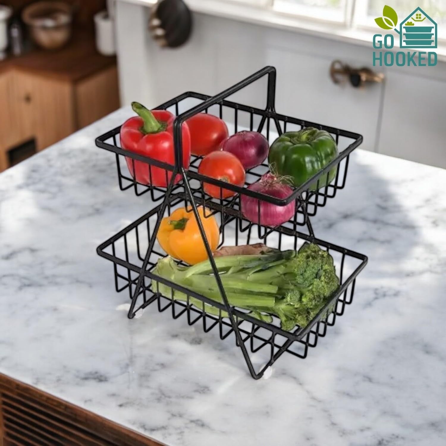GoHooked black metal 2-tier fruit basket with handle, holding various vegetables and fruits on a marble countertop.