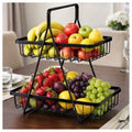 GoHooked black metal 2-tier fruit basket with handle, filled with bananas, apples, strawberries, grapes, and lemons on a dining table.
