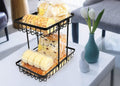 GoHooked black metal 2-tier fruit basket holding bread and pastries on a table.