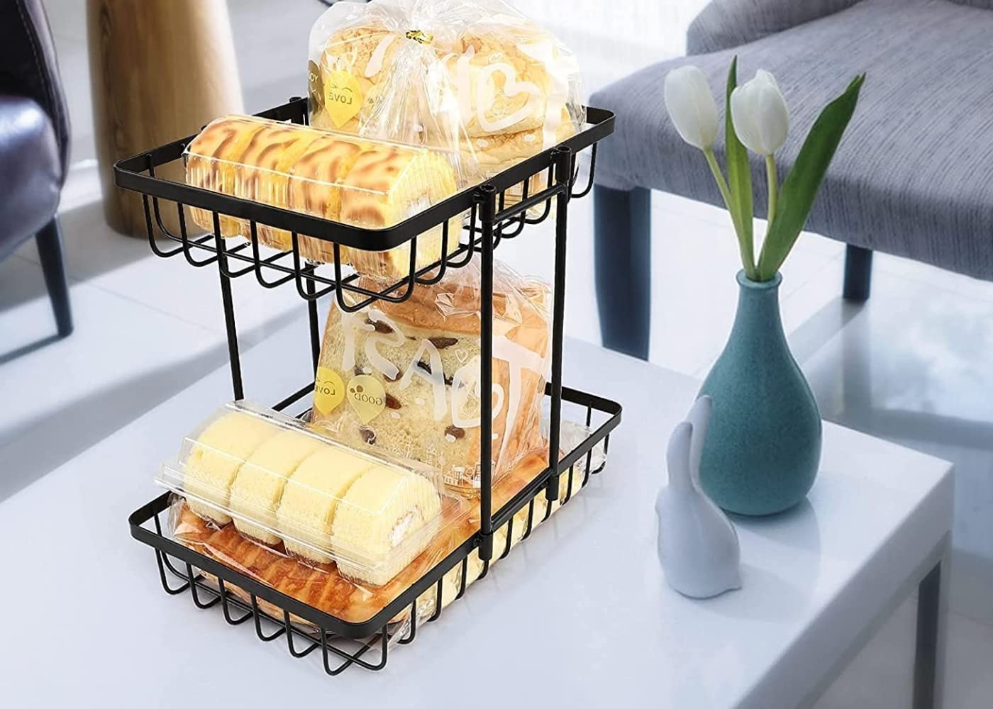 GoHooked black metal 2-tier fruit basket holding bread and pastries on a table.