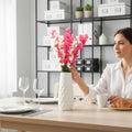 GoHooked pink and white artificial flowers in a textured white vase, a woman adjusts the bouquet on a dining table.