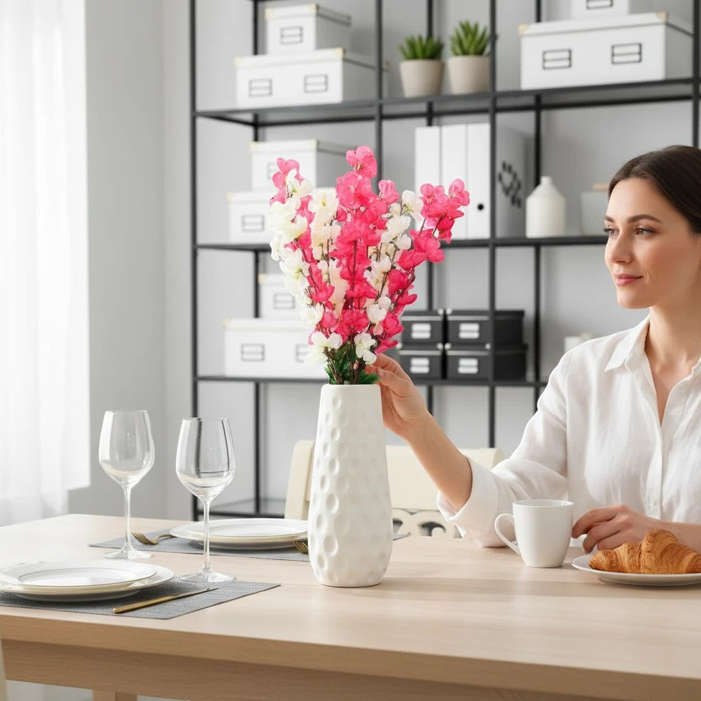 GoHooked pink and white artificial flowers in a textured white vase, a woman adjusts the bouquet on a dining table.