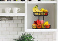 GoHooked black metal 2-tier fruit basket filled with bananas, apples, grapes, and other fruits on a kitchen counter.
