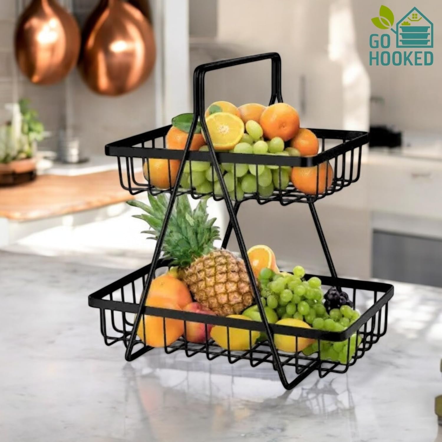 GoHooked black metal 2-tier fruit basket with handle, filled with oranges, grapes, and pineapple on a kitchen counter.
