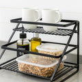 Go Hooked black metal 2-tier standing rack organizer with step shelves holding mugs, oil bottles, and food containers.