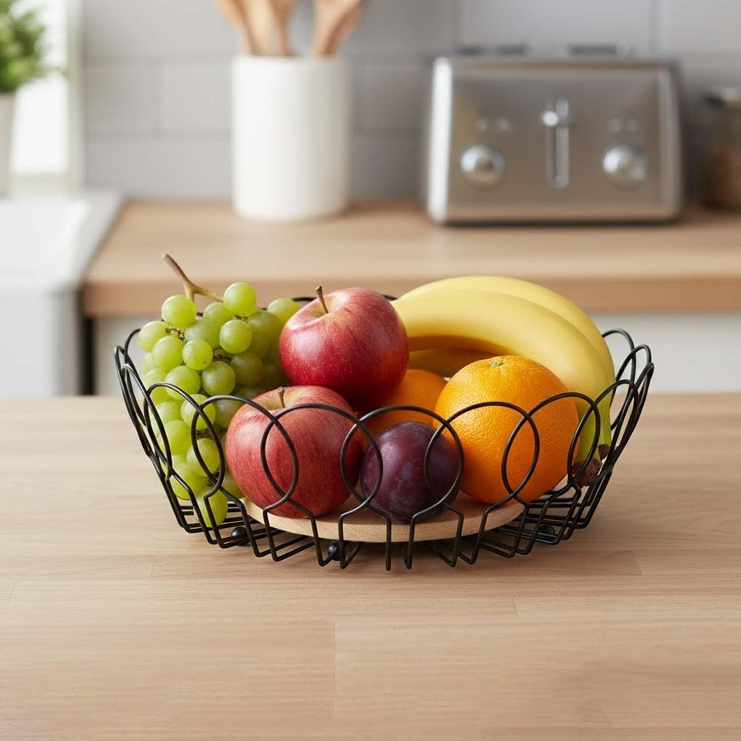 Go Hooked black metal wire fruit basket with wooden base, holding apples, bananas, oranges, plums, and grapes.