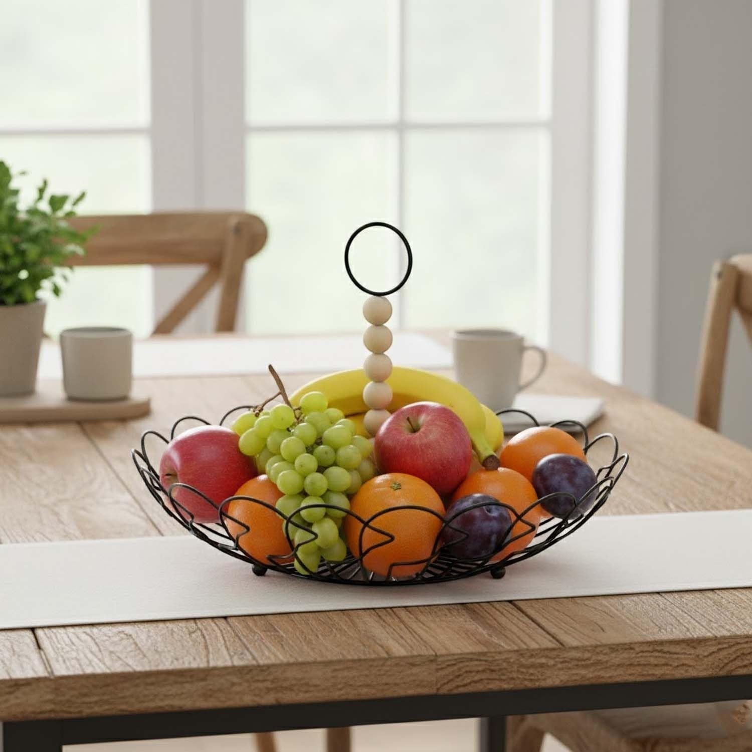 Go Hooked black metal fruit basket with decorative wire design and detachable wooden bead handle, filled with apples, oranges, grapes, bananas, and plums.