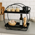 GoHooked black two-tier metal vanity storage organizer with round frame, holding jars, a mug, olive oil, and garlic.