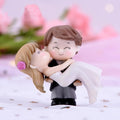 Go Hooked romantic couple figurines: groom in black suit carrying bride in white dress, bride has pink rose in hair.