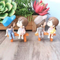 GoHooked resin couple figurines sitting on benches, one pair kissing, holding plush toys. Romantic decor.