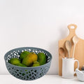 Go Hooked light grey round plastic fruit basket with decorative cutouts, holding fresh mangoes.