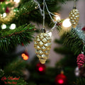 Go Hooked golden pine cone Christmas ornaments hanging on a tree with festive lights.
