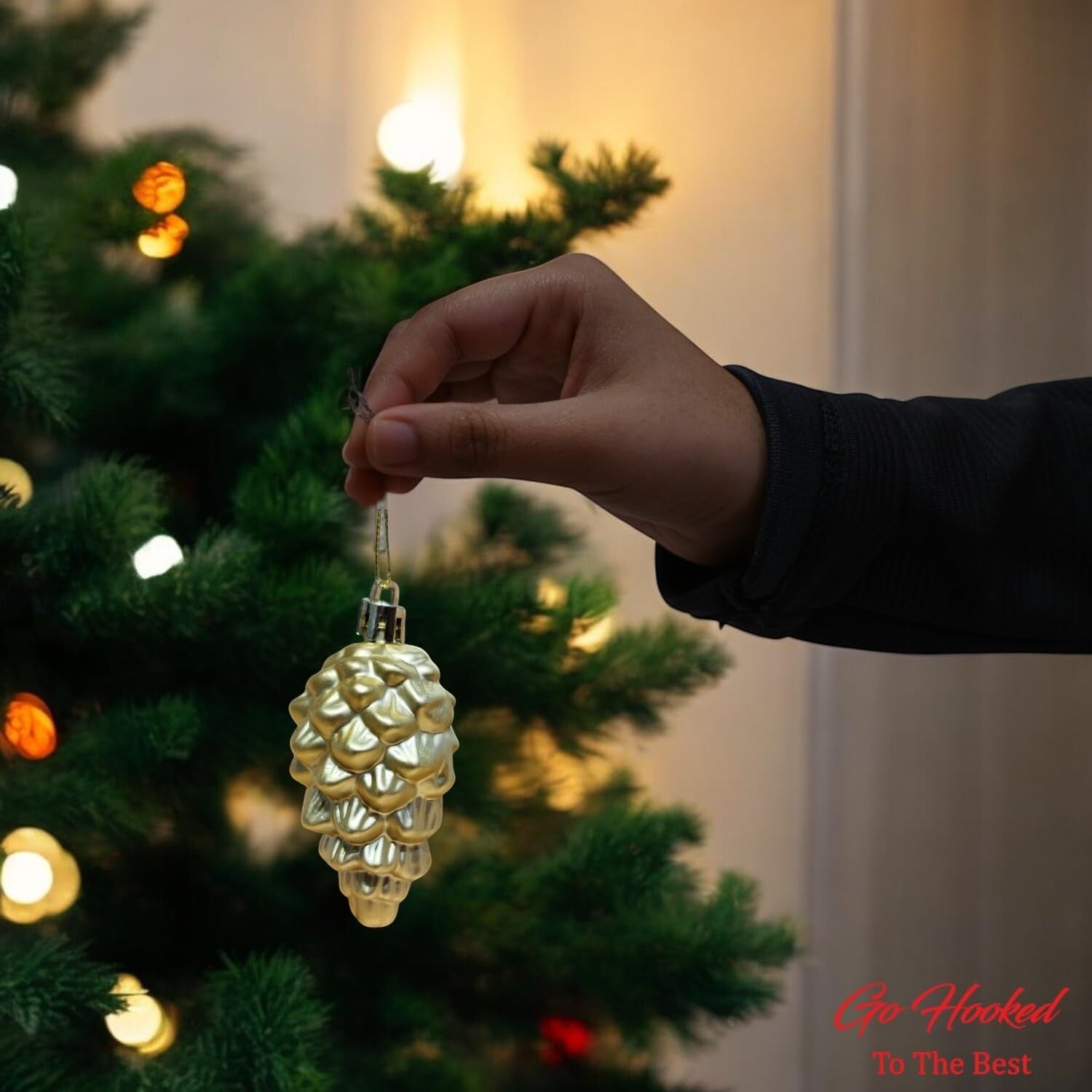 Go Hooked golden pine cone Christmas ornament with glitter details, held by a hand near a decorated tree.