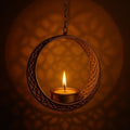 GoHooked matte gold metal hanging tealight holder with floral cut design and chain. A lit candle inside casts a warm glow.