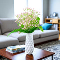 GoHooked light pink artificial baby's breath flowers in a white textured vase, with green leaves, on a coffee table.