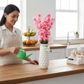 GoHooked light pink artificial flowers in a textured white vase, a woman waters them with a green watering can.