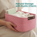 Go Hooked light pink rectangular cotton rope storage basket with brown PU leather handles, filled with folded blankets and books.