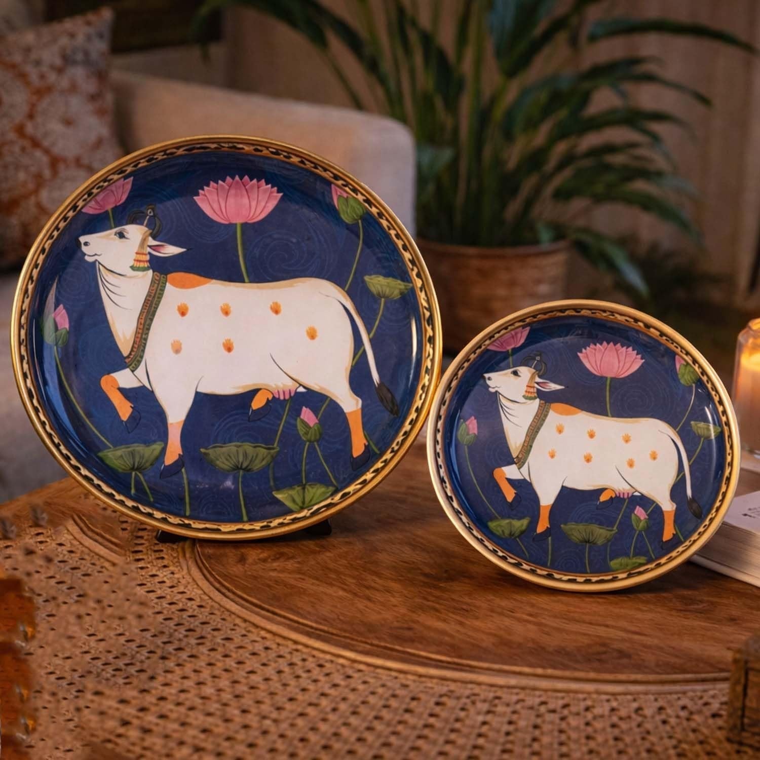 Go Hooked set of 2 round metal decorative trays with gold rims, featuring a white cow and pink lotus flowers on a dark blue background.