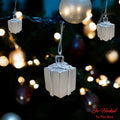 Go Hooked silver gift box Christmas ornaments with glitter accents, hanging on a decorated tree with bokeh lights.
