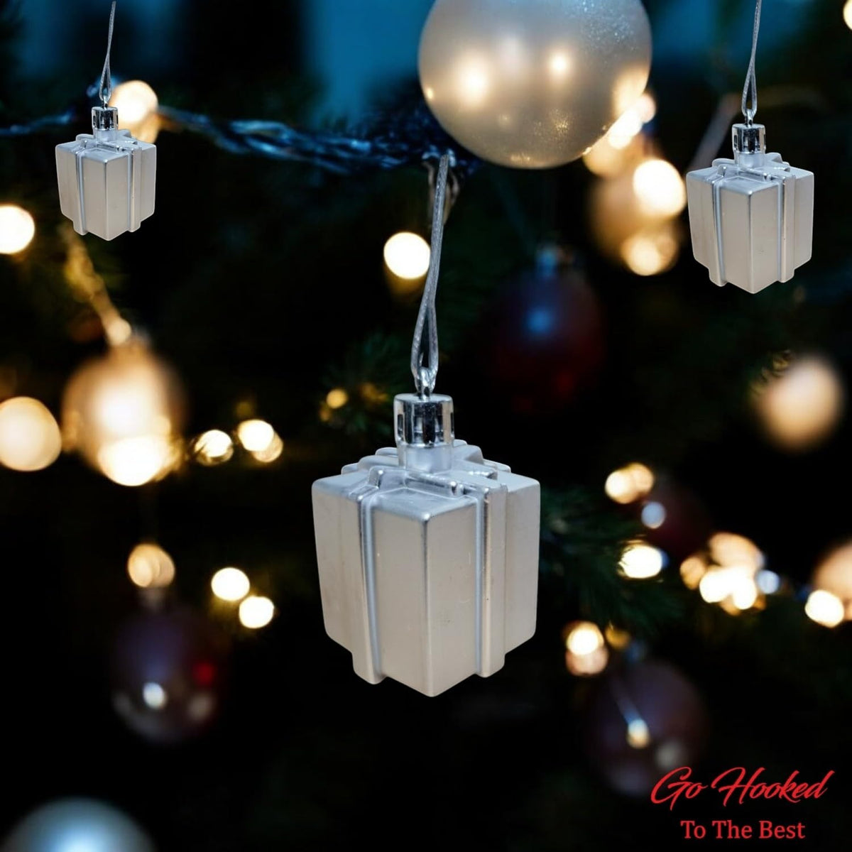 Go Hooked silver gift box Christmas ornaments with glitter accents, hanging on a decorated tree with bokeh lights.