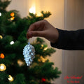 Go Hooked silver pine cone Christmas ornament with glitter details, held by a hand near a decorated tree.