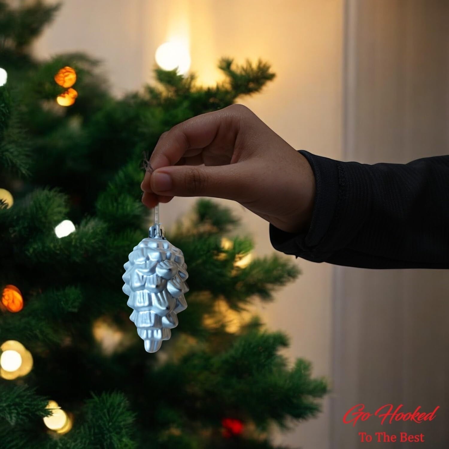 Go Hooked silver pine cone Christmas ornament with glitter details, held by a hand near a decorated tree.