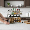 GoHooked stainless steel 2-tier shelf basket organizer filled with spice jars, condiment bottles, and pepper mills on a kitchen counter.
