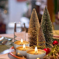 GoHooked 50pcs tealight candles with warm flickering flames, set on a table with mini bottle brush trees and red ornaments.