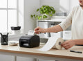 GoHooked white thermal barcode labels, 100x150mm, on a roll next to a label printer. Ideal for warehouses and shipping.