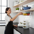 GoHooked white metal undershelf basket holding colorful bowls, with a woman reaching for a bowl.