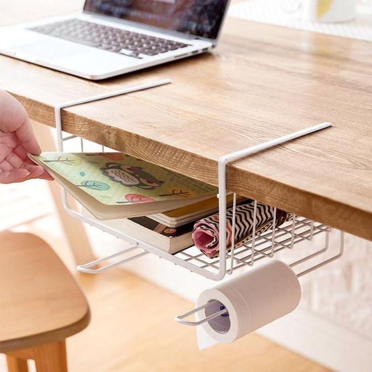 Go Hooked white metal under shelf basket holding books and a roll of paper towels, attached to a wooden desk.