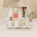 Go Hooked white 2-tier metal vanity organizer with soap dispenser, toiletries, makeup brushes, and cotton swabs.